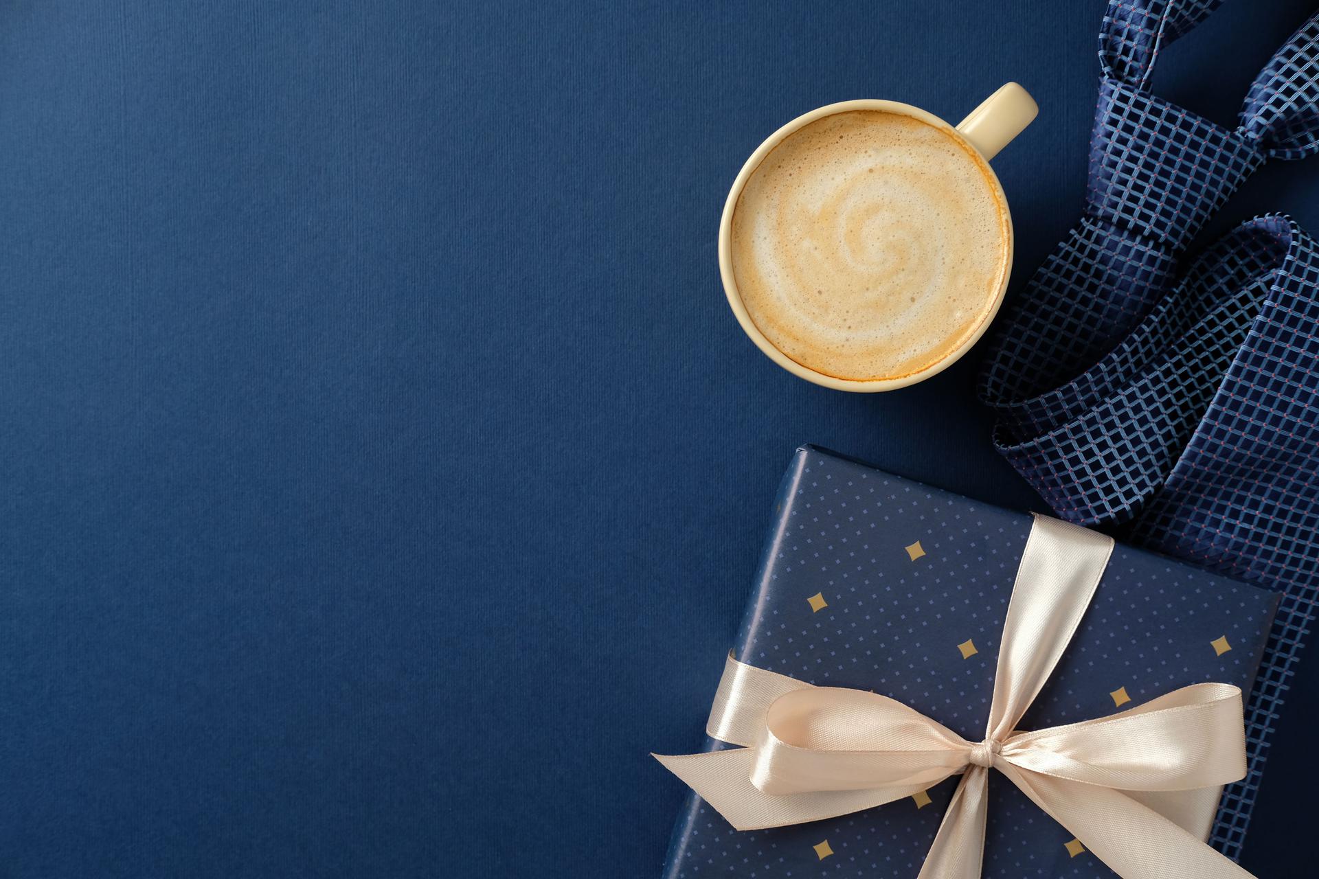 Happy Fathers day concept. Stylish flat lay of a gift box with a cream ribbon, coffee cup, and patterned blue tie on a dark blue background